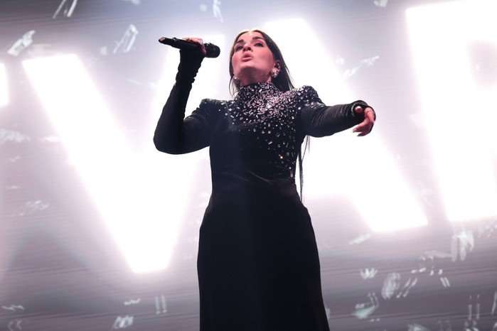 Atvara performing on stage wearing a long black sparkly gown, singing into a microphone.