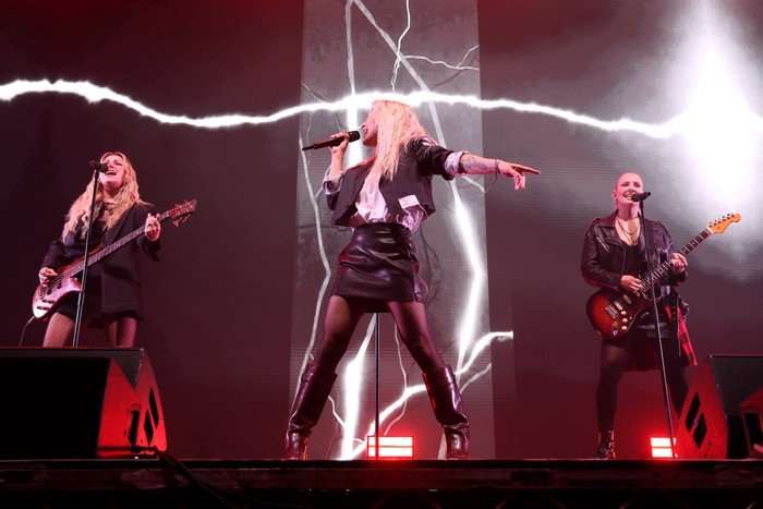 Three women stood in a line performing on stage. The two on the outside sing into microphones whilst playing guitars and the lead singer in the centre sings into a microphone with her arm stretched out.