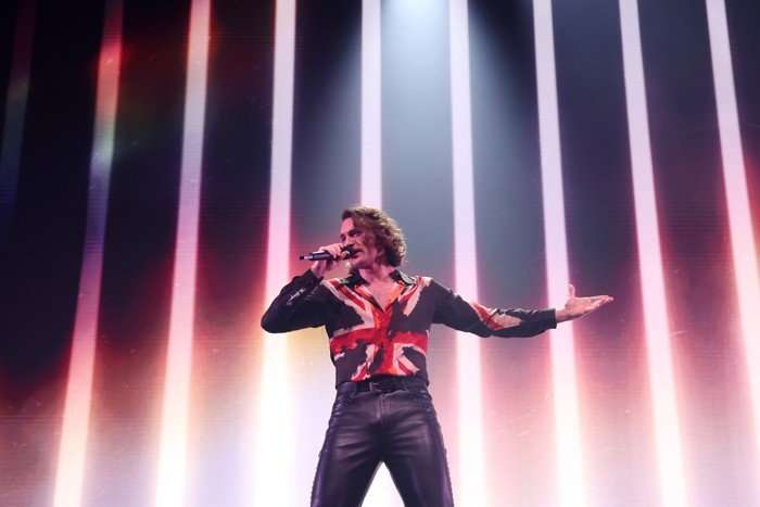 Aidan stood on stage singing into a microphone with one arm stretched out.