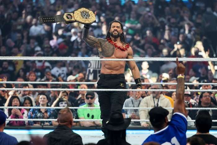WWE wrestler Roman Reigns, topless in black trousers, holds up the World Heavyweight Championship in the ring at WrestleMania 42.