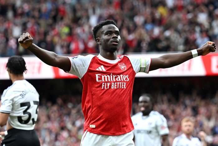 Arsenal attacker Bukayo Saka, in red and white football kit, holds both arms out wide as he stands in celebration on the pitch at the Emirates.