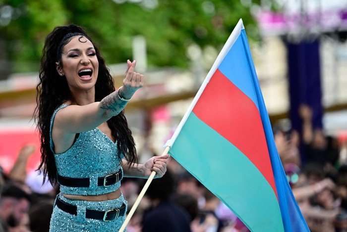 Eurovision-2026-azerbaijan Jiva smiling wide on stage holding the Azerbaijan flag and making a heart with her other hand.