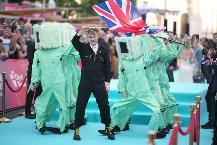 Sam Battle wearing a dark blue boiler suit walking on a light blue carpet waving a UK flag.