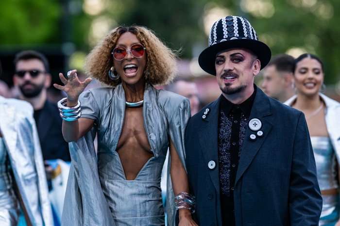 Senhit and Boy George walking hand in hand down a light blue carpet.