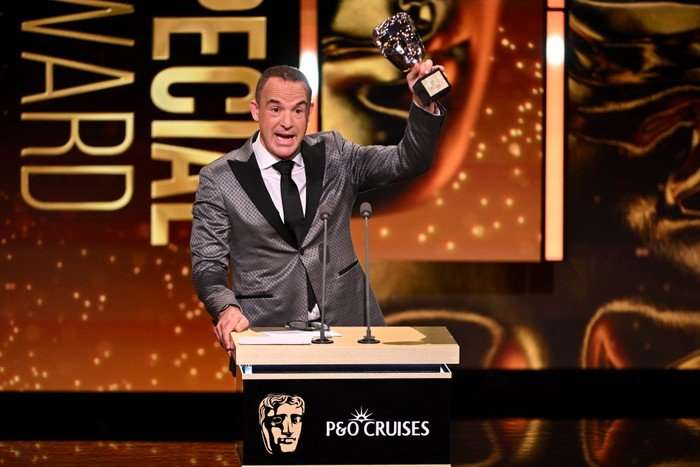 Martin Lewis in a grey suit with black lapels, holds a gold trophy in the shape of a face over his head on stage