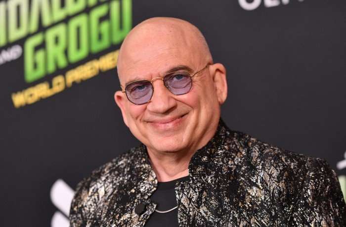 English actor Jonny Coyne attends the world premiere of "Star Wars: The Mandalorian and Grogu". He wears smart purple shades and a artistically patterned dark jacket. He smiles at the camera.