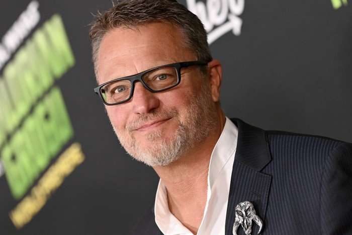 Steve Blum attends the Premiere of Disney's "Star Wars: The Mandalorian and Grogu". He wears thick-rimmed black glasses and a white shirt and dark blue suit jacket. He smiles towards the camera.