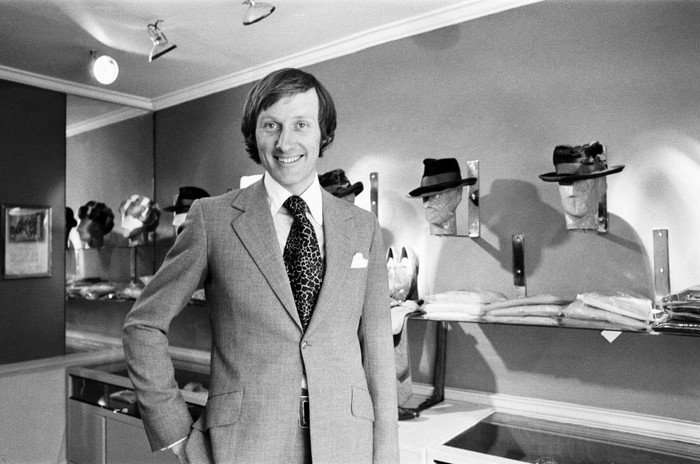 A 1969 black and white photo of Rupert Lycett wearing a suit and smiling at the camera in front of mannequins at Tailors in Savile Row
