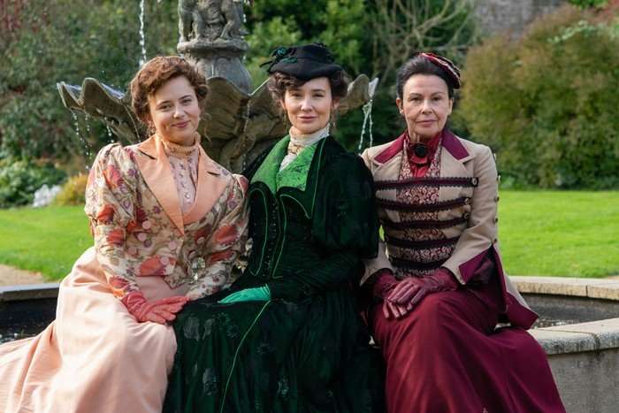 Three women sit in period dress in a garden, they are all sitting upright, posing for the cameras with their hands in their laps