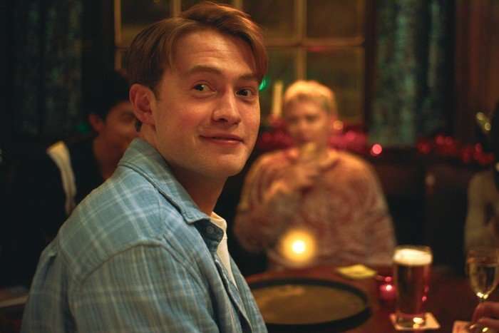 Kit Connor as Nick in Heartstopper Forever, wearing a blue shirt and looking round at someone, smiling.