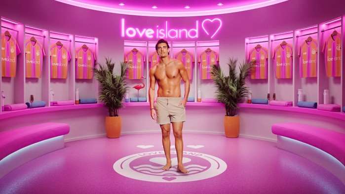 ©ITV A man stood in a pink room wearing tanned swim shorts with one hand in his pocket.