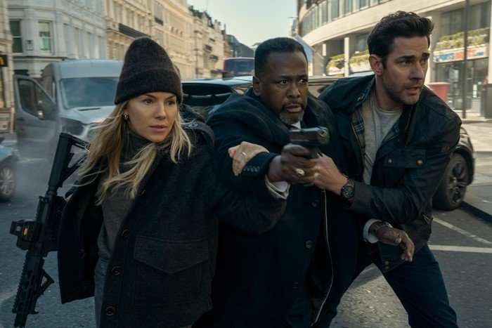 Sienna Miller as Emma Marlowe, Wendell Pierce as James Greer and John Krasinski as Jack Ryan in Tom Clancy's Jack Ryan: Ghost War. They are stood in a street, with Greer holding out a gun.