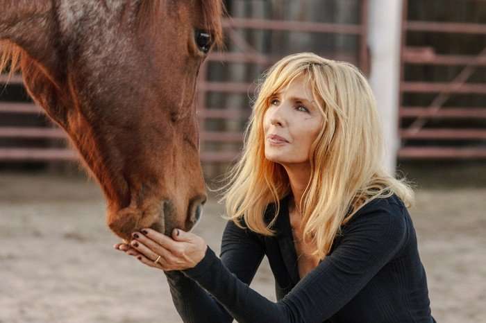 A blonde woman crouches beside a brown horse in a stable or ranch setting, gently holding the horse&rsquo;s muzzle in her hands. She looks up at the animal with a soft, affectionate expression. The woman wears a fitted dark long-sleeved top, while the background features sandy ground and red metal fencing blurred out behind them.