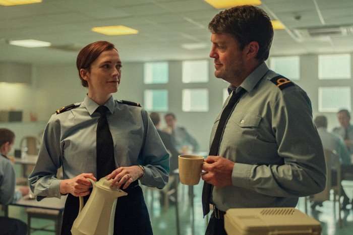 Tom Burke and Charlotte Ritchie in Legends, talking in an office, wearing customs officer uniforms. He is holding a mug while she is holding a coffee pot.
