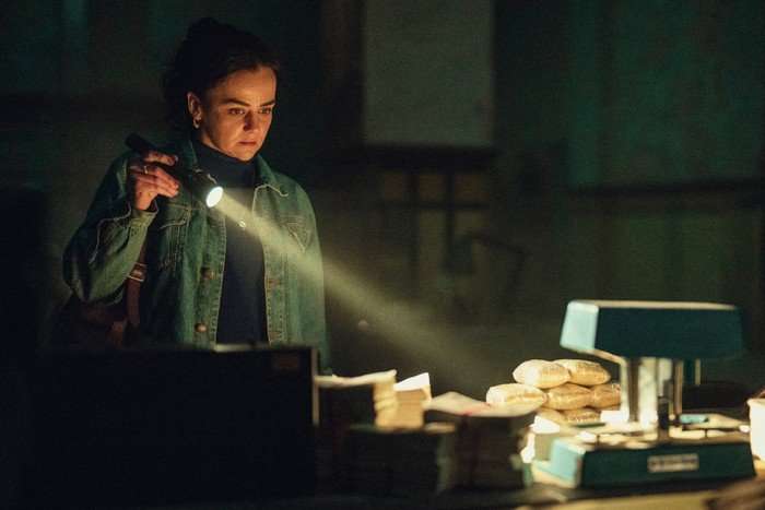 Hayley Squires in Legends, wearing a denim jackets, stood in a dark room and holding a torch. The light is beaming down a table containing packages of drugs and packets of cash.