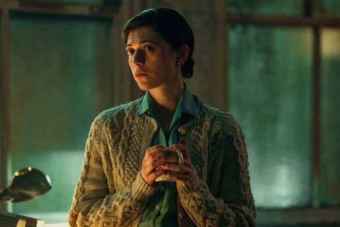 Jasmine Blackborow as Erin in Legends, wearing a shirt and cardigan and holding a mug with both hands.