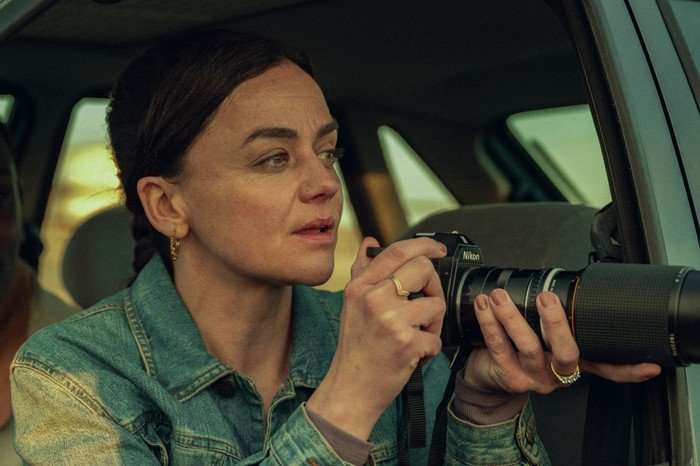 Hayley Squires in Legends, wearing a denim jacket and leaning out of a car window, holding a long-lens camera.