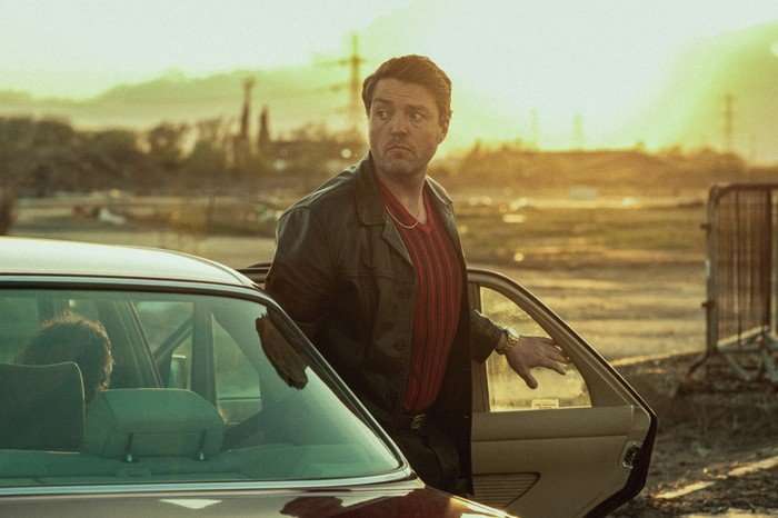 Tom Burke in Legends, wearing a red top and leather jacket and stepping out of a car door.