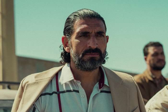 Numan Acar as Hakan in Legends, stood outside wearing a brown jacket and white shirt.