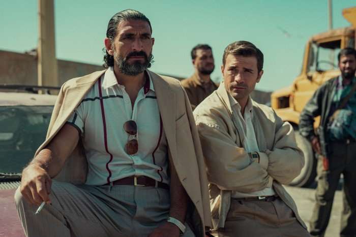 Numan Acar as Hakan and Kem Hassan as Aziz in Legends, sat on a car in a sunny location, with others stood behind them.