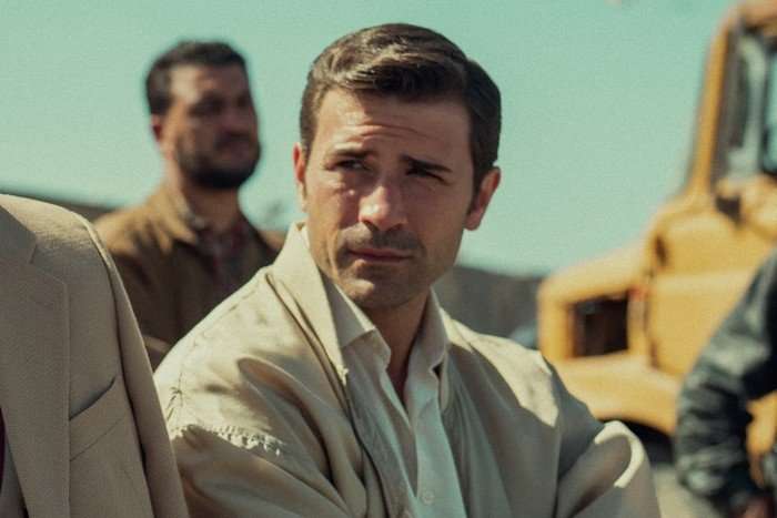 Kem Hassan as Aziz, in Legends. He is outside wearing a brown jacket and white shirt. There is another man stood behind him.