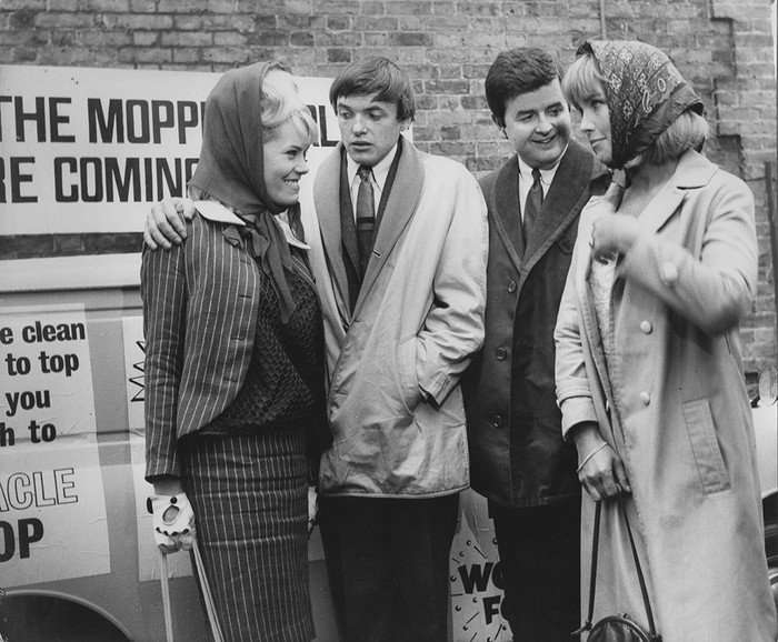 Likely Lads S2E4 Big Spenders 05 A black-and-white still from The Likely Lads, featuring James Bolam as Terry Collier and Rodney Bewes as Bob Ferris standing with two women, all dressed in mid-1960s attire. The group is pictured smiling and chatting near a van with a sign reading "The Moppetts are coming," capturing a lighthearted moment typical of the show's humour.