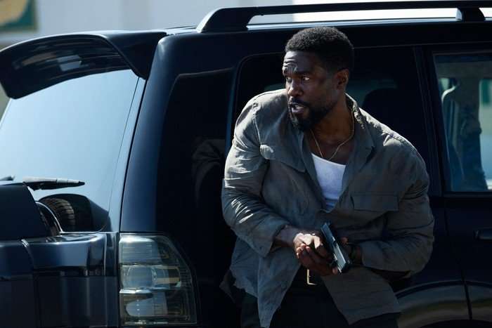 Show moreShow lessA man steps out of the back of a dark SUV, holding a handgun in both hands and scanning his surroundings with a tense, alert expression. He wears a partially unbuttoned shirt over a white vest, and the scene suggests a moment of imminent danger or action.
