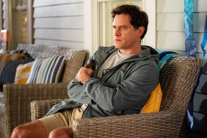 THE FOUR SEASONS, SEASON 2 A man sits in a wicker chair on a porch or patio, holding a bottle close to his chest with his arms folded. He is wearing a grey T-shirt, beige shorts and a light waterproof jacket, and looks ahead with a serious, contemplative expression. Cushioned outdoor seating and blue-and-yellow decorations surround him in the softly lit setting.