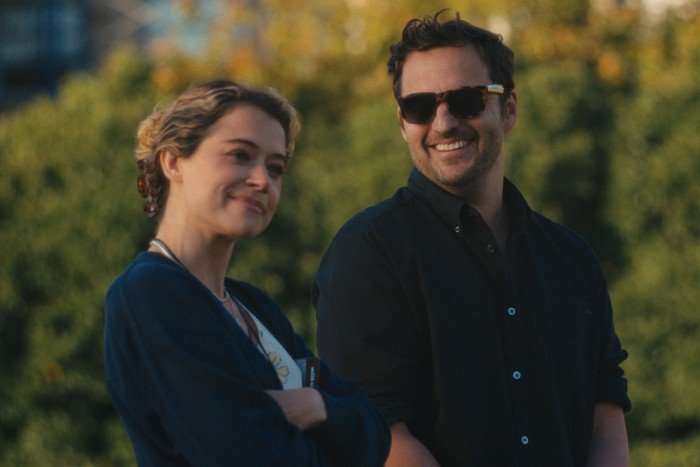 Tatiana Maslany and Jake Johnson in Maximum Pleasure Guaranteed, stood next to each other outside. She is smiling ahead while he is wearing sunglasses and looking at her, smiling.