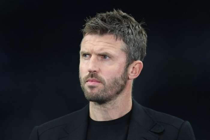 Michael Carrick, in a black t-shirt and black suit jacket, stares intently in front of a dark background.