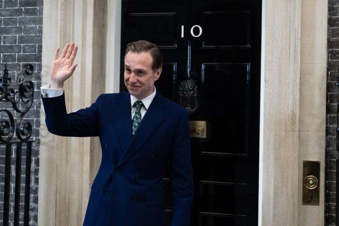 Rafe Spall as Prime Minister Harry Douglas in Number 10, wearing a suit, stood outside 10 Downing Street, waving.