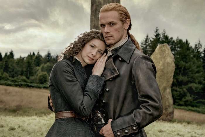A couple in period clothing stand embracing in a field beside standing stones, with dark clouds gathering overhead.