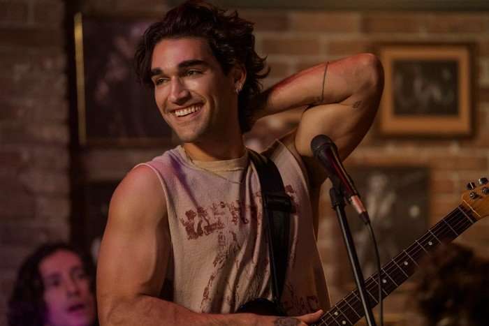 Josh Heuston as Justin in Off Campus; in this scene, he is stood on a stage in front of a microphone, wearing a guitar around his neck and flexing his bicep as he smiles at the crowd