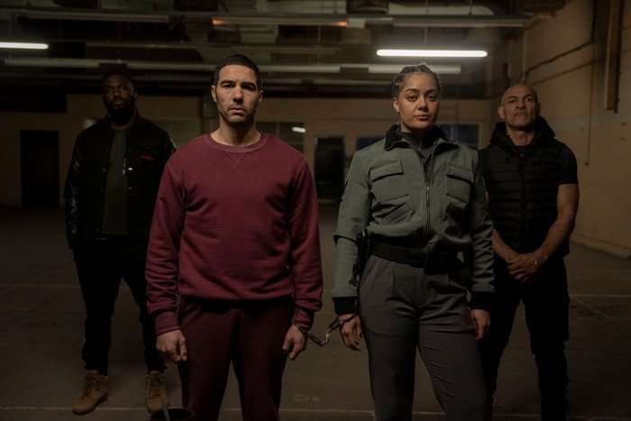 Prisoner Izuka Hoyle and Tahar Rahim in Prisoner standing in an underground office, being flanked by other people while standing in the middle of the room cuffed together.