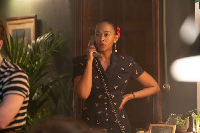 A woman stands indoors speaking on a corded telephone, looking concerned as she rests one hand on her hip. She is wearing a dark short-sleeved dress with gold detailing, statement earrings and a red hair accessory. Warm lighting, wooden panelled walls and houseplants can be seen around her, while blurred figures appear in the foreground.