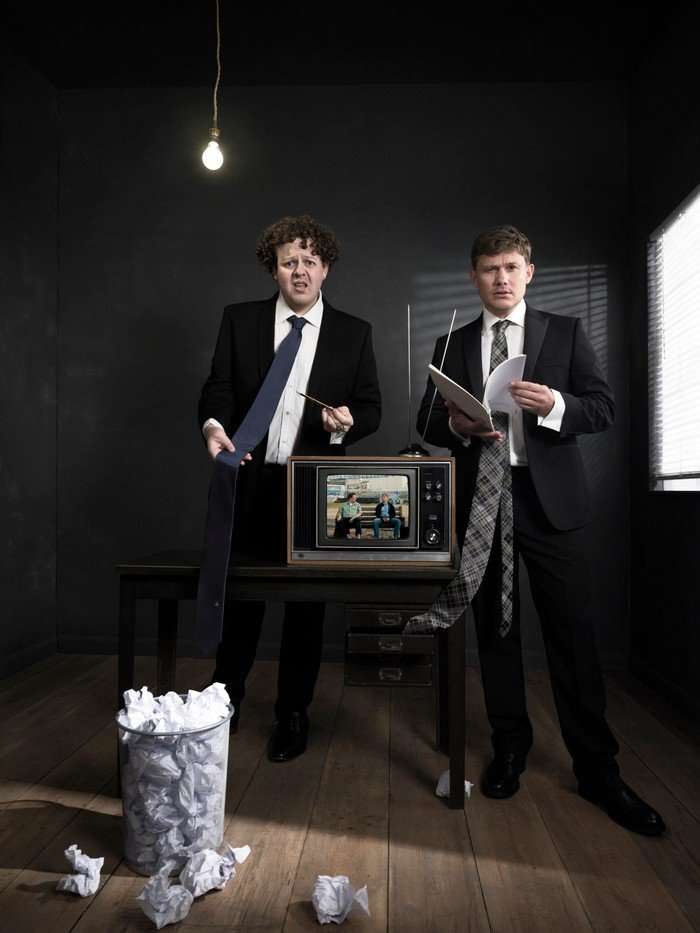 Jack Rooke and Jon Pointing wearing black suits in a dark room with a wastebin full of paper in front of them.