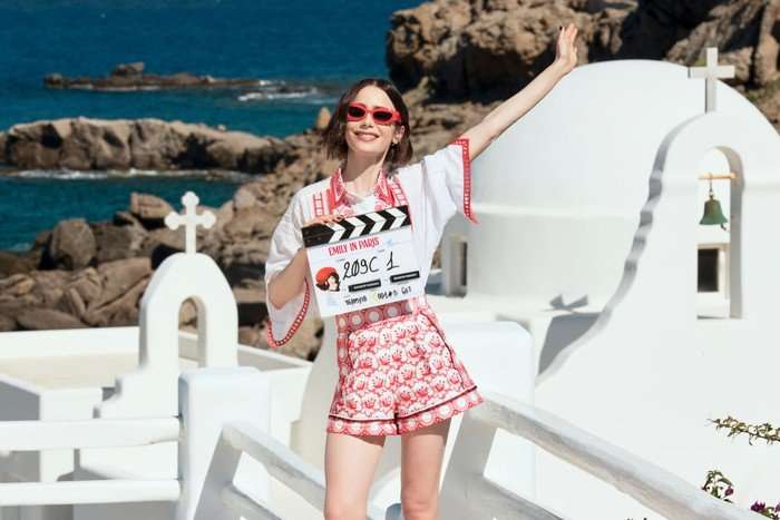 Emily in Paris Production Image season 6 Lily Collins poses with a clapperboard in a pair of red shorts at a lookoutpoint in Santorini, Greece