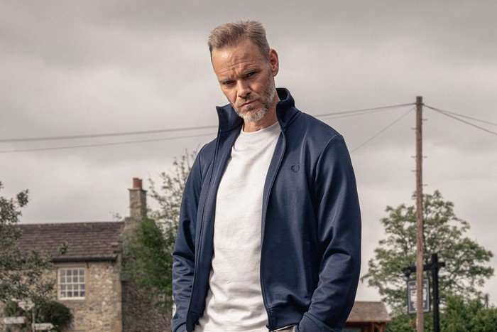 Ray (Joe Absolom) stands with his hands in his pockets in a blue jacket, white t-shirt and grey trousers