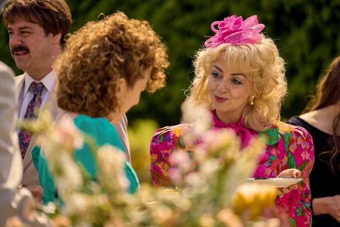 Lisa McGrillis as Valerie Jones in Rivals wearing a pink hair fastening and chatting to Katherine Parkinson in a garden.