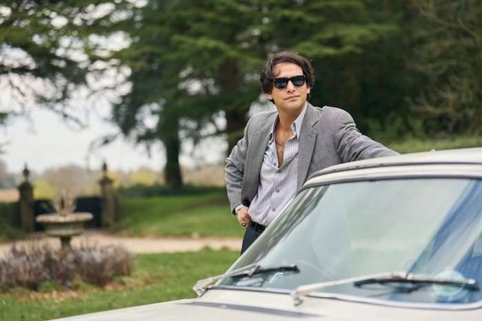 Luke Pasqualino as Bas Baddingham in Rivals wearing a suit and sunglasses while resting on the door of his car.