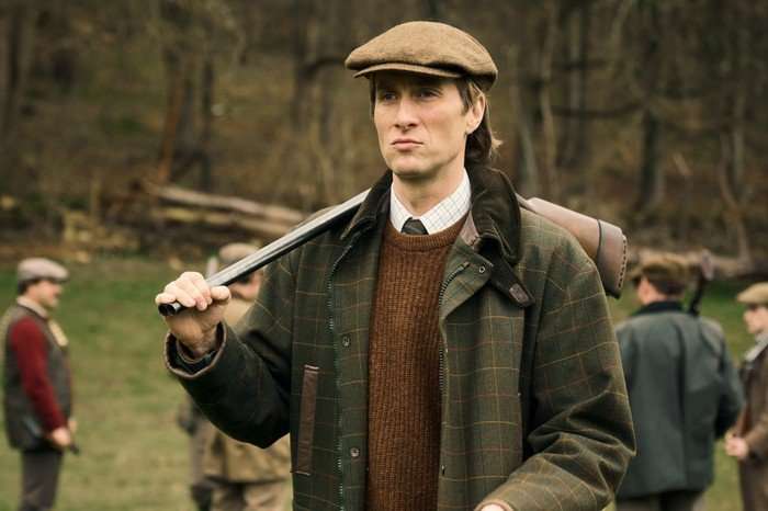 Oliver Chris as James Vereker in Rivals holding a rifle over his shoulder and wearing a hat and coat while walking in a field.