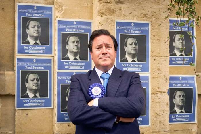 Rufus Jones wearing a suit, blue tie and ribbon, standing in front of posters of himself and crossing his arms.