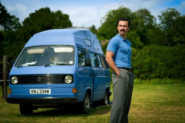Aidan Turner as Declan O'Hara standing with his hands in the pockets of his trousers, wearing a bright blue polo t-shirt and standing in a field in front of a blue camper van.