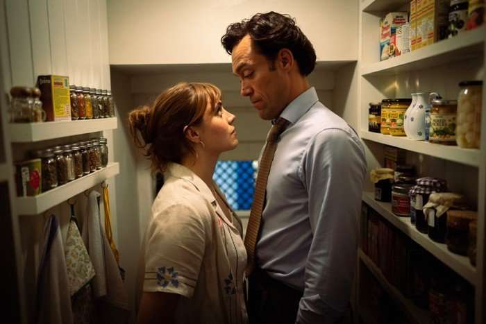 Bella Maclean and Alex Hassell standing face to face and close together as they stand in a pantry, with Bella wearing an apron and Alex wearing a shirt and tie.