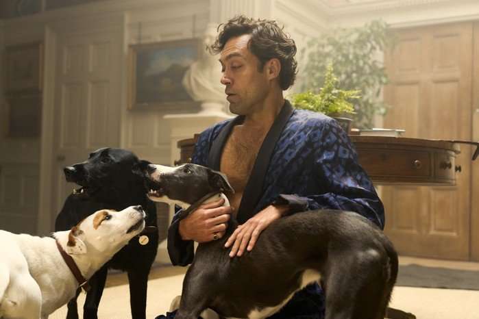 Alex Hassell sitting on the floor, surrounded by dogs and smiling as they nuzzle each other.