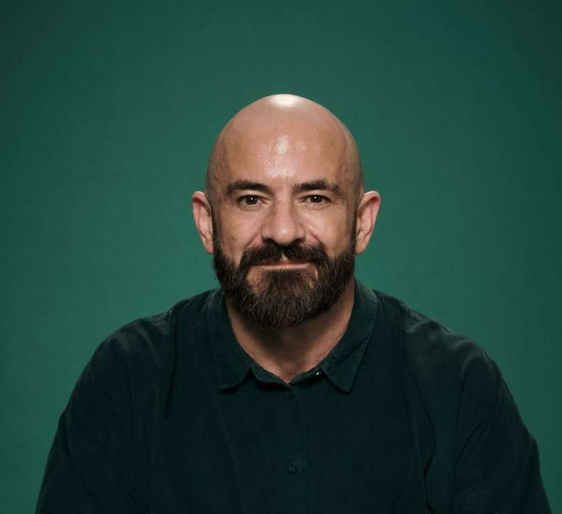 A bearded Gareth McLean sits before a dark green backgrounded, looking at the camera and smiling gently