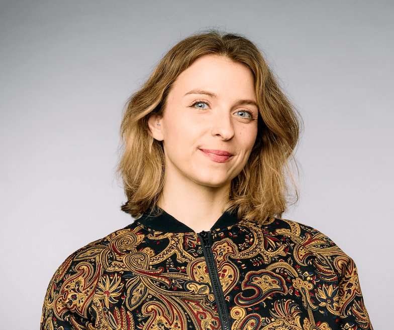 A head and shoulders shot of Abby Robinson. She is in front of a grey background, looking at the camera and smiling. She wears a black zipped-up jacket with an elaborate gold and maroon design