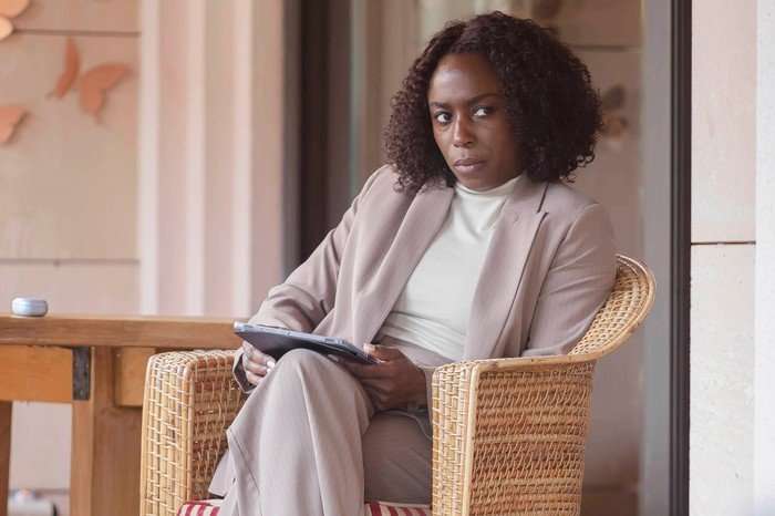 A woman sits in a wicker chair on what looks like a patio or outdoor seating area. She&rsquo;s wearing a light beige suit with a high-neck top underneath and holds a tablet in her hands. Her posture is relaxed but slightly angled, with one leg crossed over the other. She has shoulder-length curly hair and is looking off to the side with a serious, thoughtful expression&mdash;almost as if she&rsquo;s observing someone or listening carefully. The setting is softly lit and calm, with neutral tones, a wooden table nearby, and subtle decorative elements in the background.