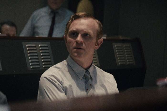 Josef Davies as Sergei Nikulov in Star City, wearing a grey shirt and tie and sat at a desk.