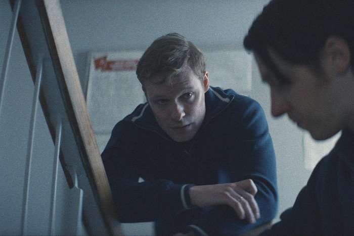Solly McLeod as Sasha Polivanov in Star City, leaning on a desk, talking to someone sat at it. He is wearing a blue top.